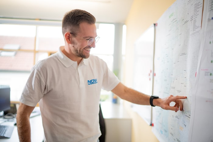 Mann in weißem NOV-Poloshirt zeigt lächelnd auf ein Whiteboard mit Diagrammen.